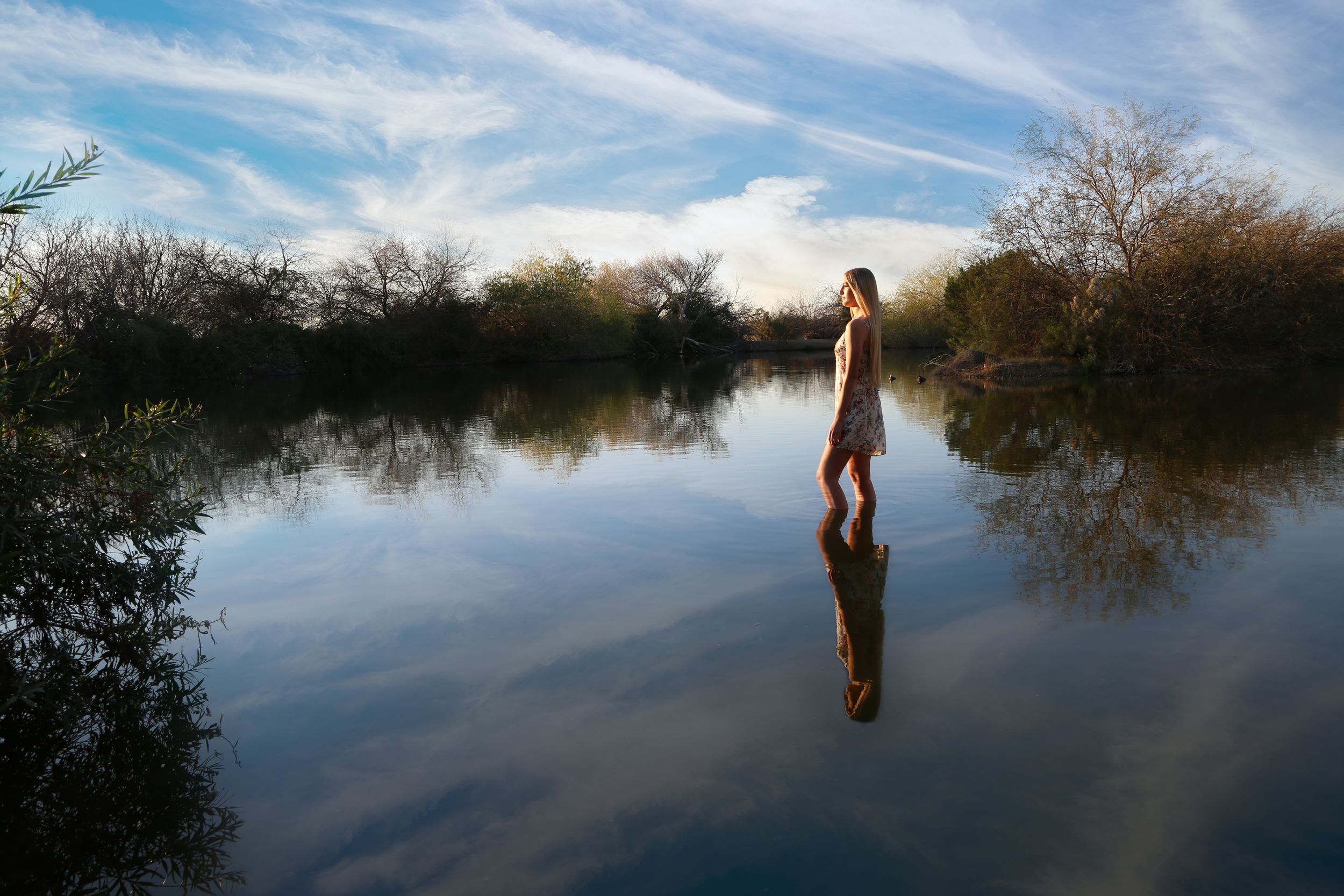 Woman standing in a calm lake with her reflection in the water, surrounded by trees under a blue sky with clouds.