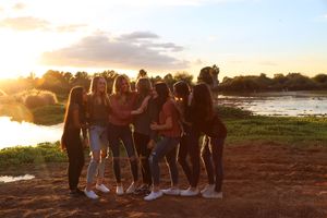 Group of young women standing together with arms around each other by a lake at golden hour sunset.