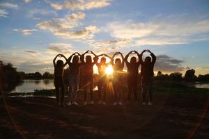 Silhouetted group of friends forming heart shapes with their arms against a bright sunset beside a lake.