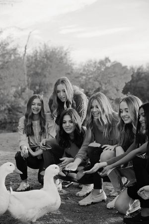 Black and white photo of smiling girls crouching outdoors to feed two white ducks by trees.
