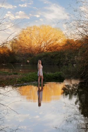 Young woman in a summer dress stands in shallow lake water reflecting golden autumn trees at sunset.