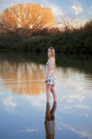 Blonde young woman in a floral dress stands in still lake water with autumn trees and clouds reflected.