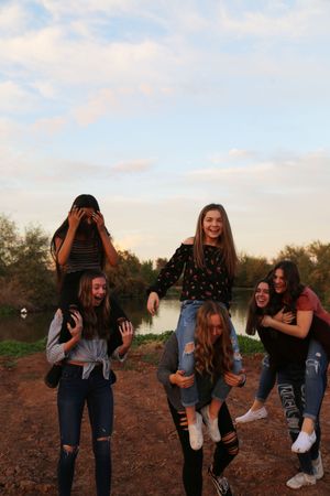 Group of laughing girls give piggyback rides on a dirt shore by a lake at dusk.