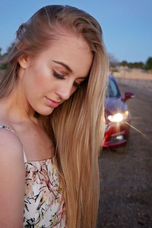Close-up portrait of a blonde young woman in a floral top looking down, with a red car behind her at dusk.