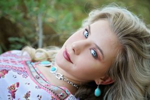 Close-up of a blue-eyed blonde woman in turquoise jewelry and a floral top lying on the ground outdoors.