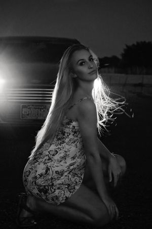 Black and white photo of a blonde woman in a floral dress kneeling before backlit vehicle headlights at night.
