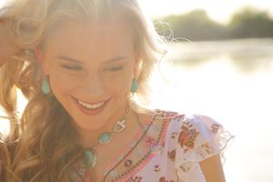 Smiling woman with long wavy blonde hair and turquoise jewelry in a floral top, bathed in warm golden sunlight by the water.