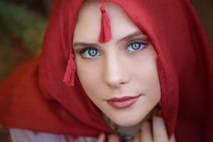 Close-up portrait of a young woman with blue eyes wearing a red hooded scarf with tassels against a soft green background.
