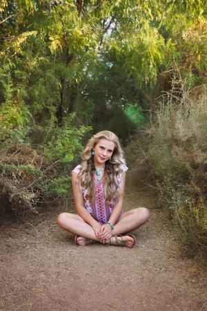 Young woman with long wavy blonde hair and turquoise jewelry sits cross-legged on a dirt path surrounded by green trees.