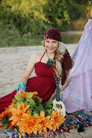 Smiling woman in a red dress and patterned headscarf with turquoise bracelets sits by orange flowers and a small ukulele.