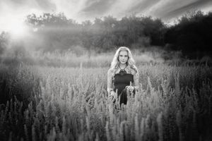 Black and white portrait of a woman with long hair standing in a tall grass field with sunlight breaking through the trees.