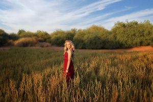 A blonde woman in a long red dress stands in a golden wheat field with green trees and a blue sky behind her.