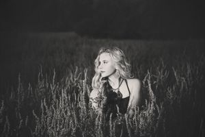 Black and white portrait of a blonde woman in a dark top sitting among tall wild grass in a moody field.