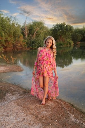 Barefoot woman in a flowing pink floral dress stands at the edge of a calm lake under a colorful sunset sky.