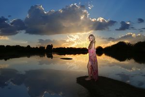 Woman in a pink dress stands on a lake shore as dramatic sunset clouds reflect across the still water.