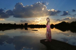 A woman in a pink dress stands barefoot on a rock beside reflective water under a dramatic sunset sky and glowing clouds.