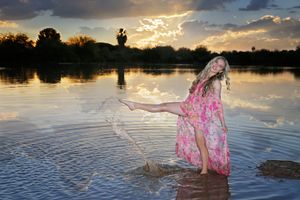 Woman in a flowing pink floral dress kicks her leg and splashes water in a lake at sunset with trees in the background.