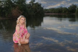 Blonde woman in a pink floral dress crouches in shallow lake water with trees and reflected clouds in the background.