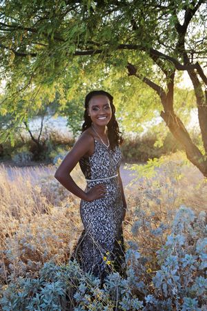 Smiling woman in a sparkly fitted gown poses with her hand on her hip beneath a green tree beside desert shrubs.