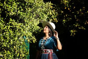 Woman in a wide-brimmed hat and turquoise top with a silver concho belt stands beside lush green citrus leaves.