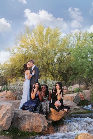 Couple kisses on a boulder while three women in formal dresses react playfully beside a small stream and blooming trees.