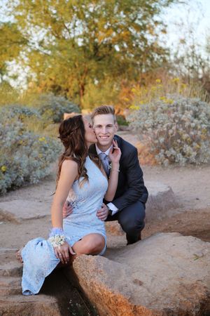 Man in a dark suit and woman in a pale blue lace dress share a kiss while posing on large rocks in a desert garden.