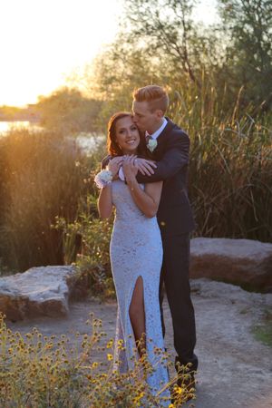 Man in a dark suit embraces and kisses a smiling woman in a light blue lace gown with a slit amid golden sunset light.