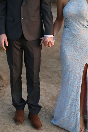 Close-up of a couple holding hands, the man in a suit and brown boots, the woman in a pale blue lace gown with a slit.