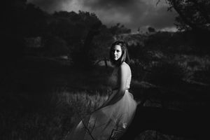 Black and white portrait of a woman in a flowing tulle skirt seated on a grassy hillside beneath a dramatic stormy sky.