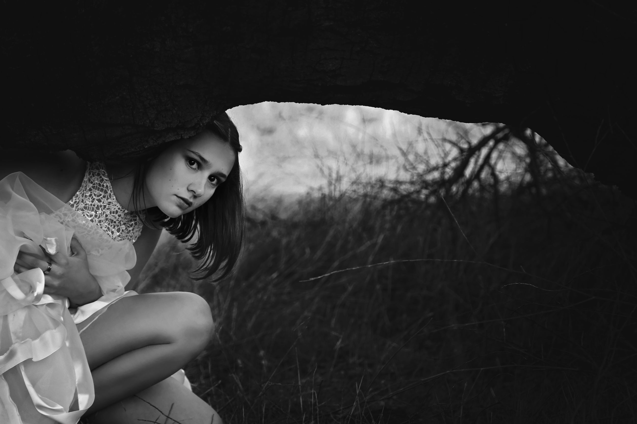 Moody black and white portrait of a young woman in a sparkly top crouching under a dark rock overhang near dry brush.