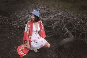 Woman in a blue hat, red floral kimono and white sundress crouches among twisted dead branches on dark ground.