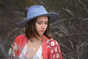 Close-up portrait of a young woman in a blue wide-brim hat and red patterned kimono looking down among bare branches.