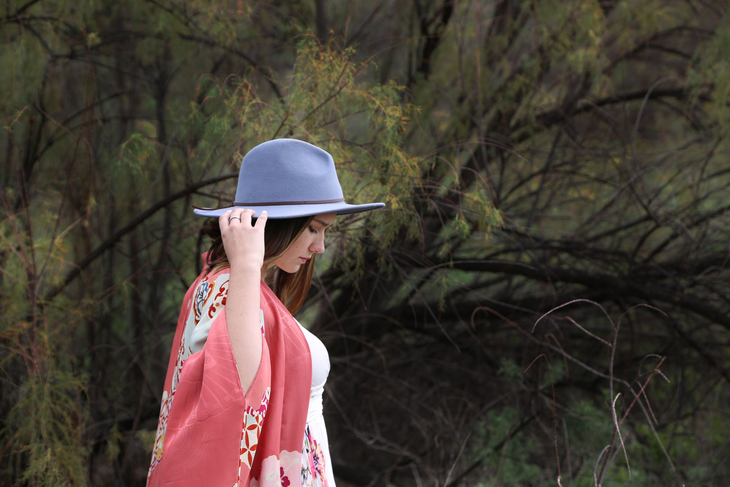 Woman in a gray-blue hat and coral kimono holds her hat brim while facing away in front of green wispy trees.