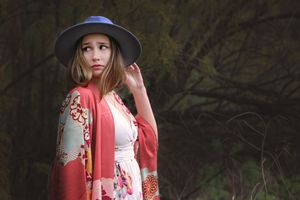 Woman in a navy hat and red floral kimono glances over her shoulder with a hand raised near dark leafy branches.