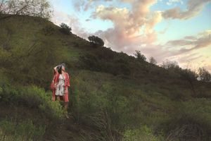 Woman in a red kimono and white dress stands on a green hillside with hands near her head under a pastel cloudy sky.