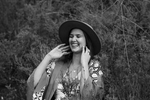 Black and white portrait of a laughing woman in a wide-brim hat and patterned kimono touching her hair among shrubs.