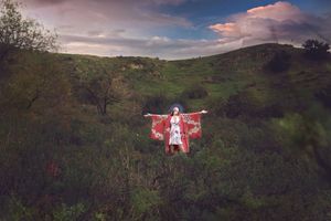 Woman in a red kimono stands with arms outstretched in a green hilly field beneath colorful sunset clouds.