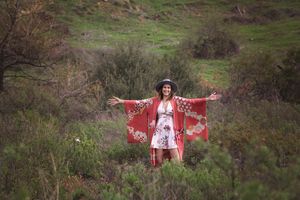 Smiling woman in a red floral kimono and hat stands with arms outstretched in a green hillside field of shrubs.