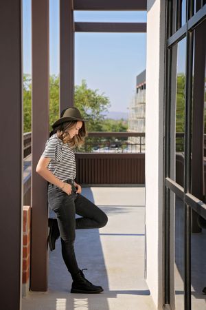 Woman in a brimmed hat, striped tee and black jeans leans against a pillar on a sunlit balcony overlooking trees and city buildings.