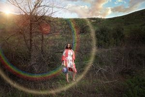 Woman in a red kimono stands on a grassy hillside holding a hat as a rainbow lens flare circles her at sunset.