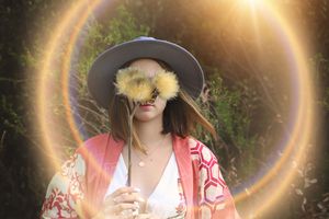 Woman in a gray hat and red kimono holds a fluffy yellow thistle flower over her face, framed by a glowing golden lens flare.
