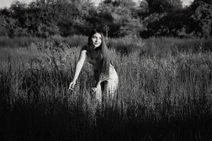 Black and white portrait of a long-haired woman leaning forward in tall grass, softly lit against a dark, shadowy field.