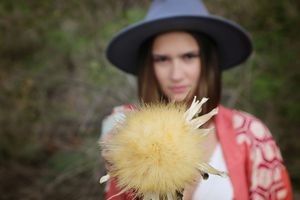 Woman in a gray hat and red patterned kimono holds a large yellow thistle flower in sharp focus against a blurred green background.