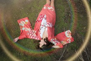 Woman in a coral floral kimono lies on her back in the grass with arms spread, encircled by a rainbow lens flare.