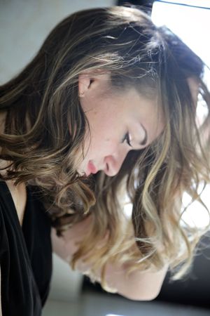 Woman with wavy, honey-highlighted brown hair looks down in profile, wearing a black top in soft natural window light.
