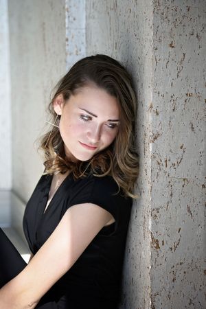 Young woman in a black top leans against a rustic textured wall, gazing to the side in dappled natural sunlight.