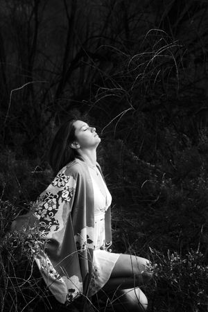 Black-and-white image of a woman in a floral kimono kneeling in dark wild grass, her face tilted skyward with eyes closed.