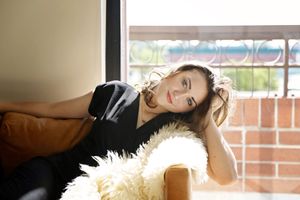 Woman in a black top reclines on a tan couch with a fluffy sheepskin throw, smiling softly near a window with brick views.