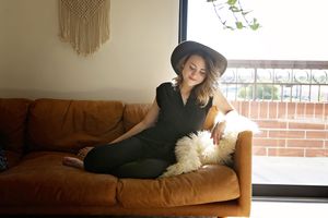 Woman in a black hat and black outfit sits cross-legged on a tan leather sofa with a sheepskin throw beside a bright window.