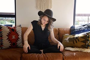 Woman in a black felt hat and black outfit sits cross-legged on a leather couch with colorful patterned pillows and macrame decor.
